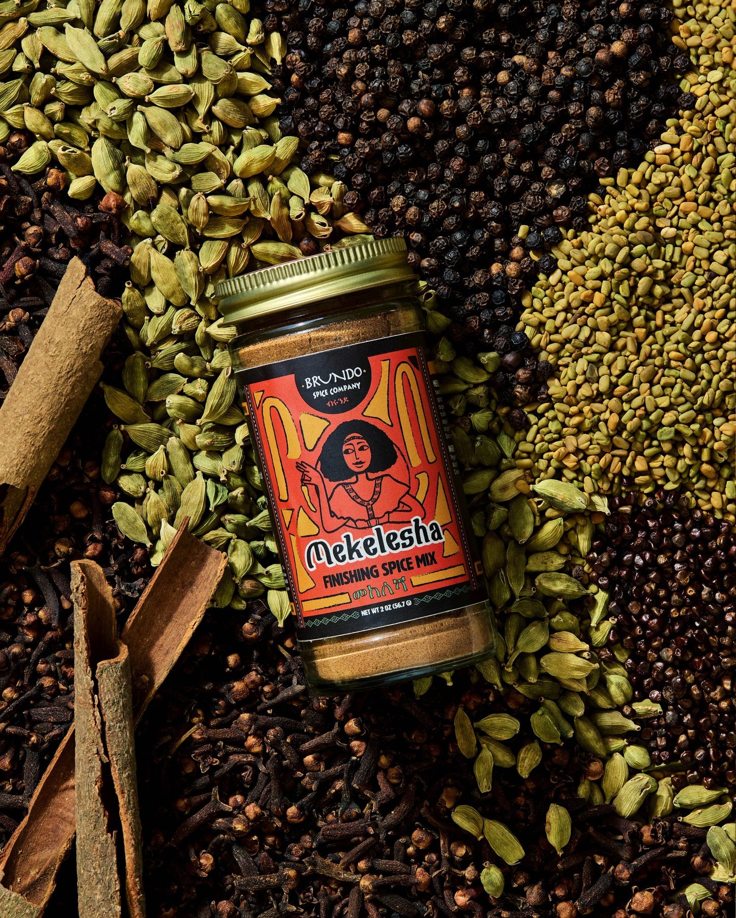 Brundo Spice Company Mekelesha Finishing Spice Mix jar surrounded by green cardamom, cloves, cinnamon bark, black pepper, and fenugreek.
