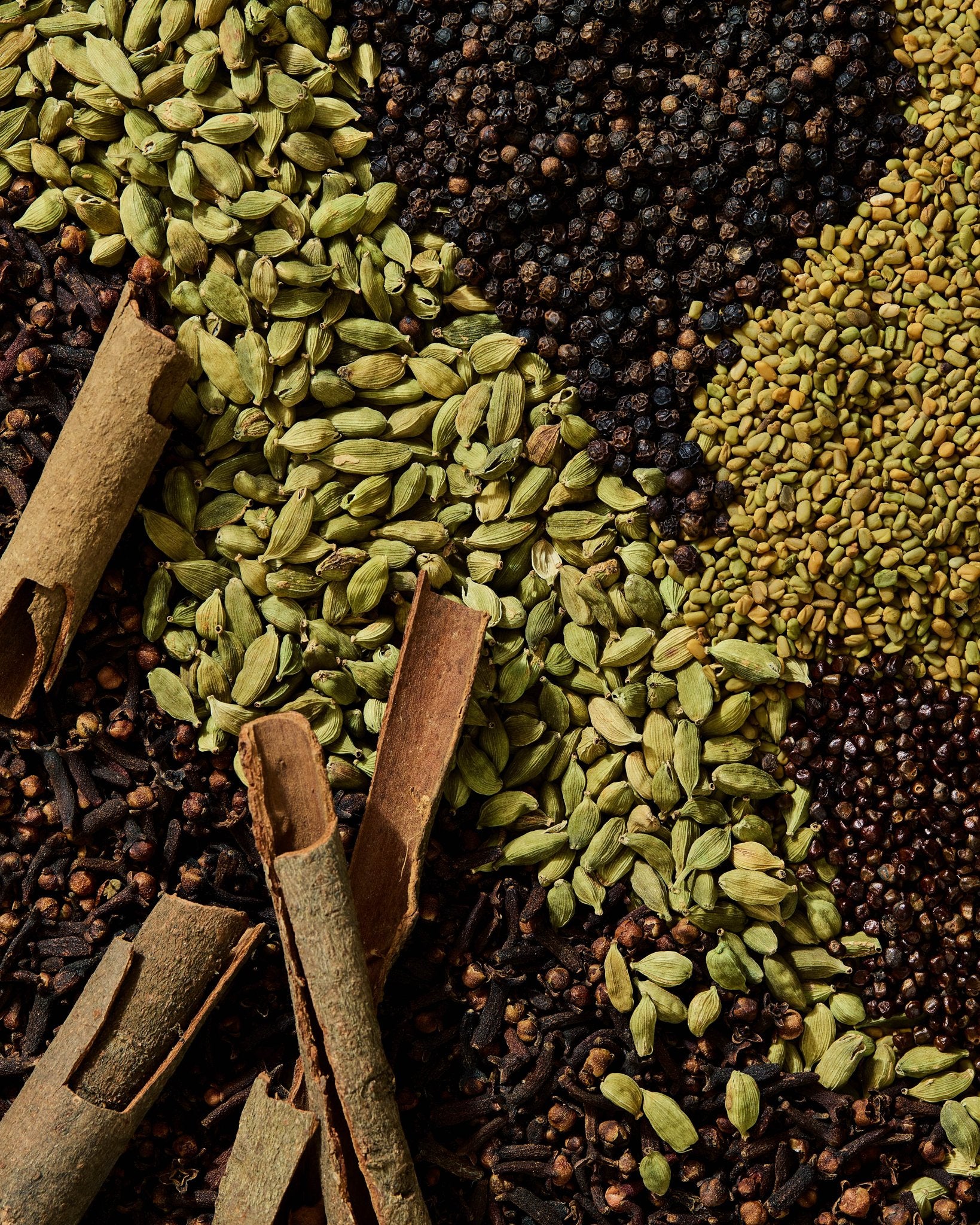 Flat lay of whole spices used in Mekelesha, including green cardamom, black pepper, cloves, cinnamon bark, and fenugreek seeds.