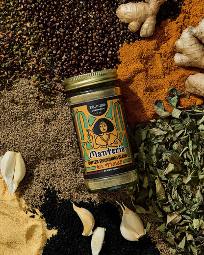 Brundo Spice Company Manteria Butter Seasoning Blend jar surrounded by ginger, garlic, turmeric, black cardamom, ajwain, and dried herbs.