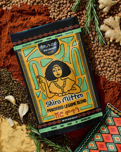 Brundo Spice Company Shiro Mitten Powdered Legume Blend pouch displayed with garlic, rosemary, chickpeas, ginger, and a traditional napkin.