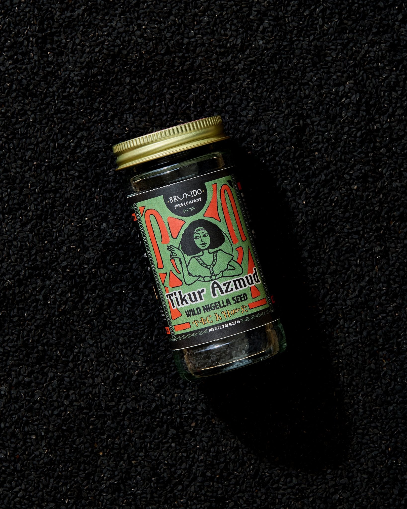Brundo Spice Company Tikur Azmud Wild Nigella Seed jar placed on a dark bed of whole black nigella seeds.